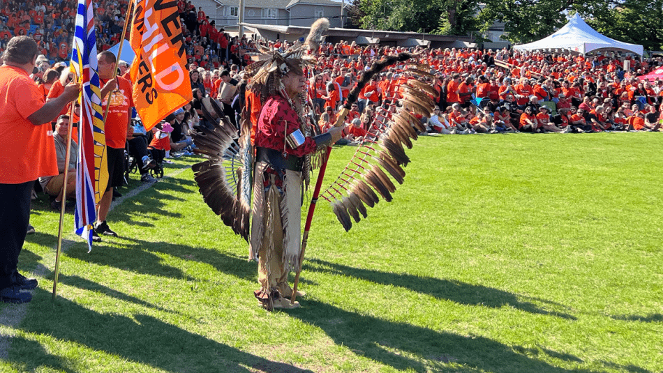 Songhees Elder leading a pow wow in front of a large crowd outside.