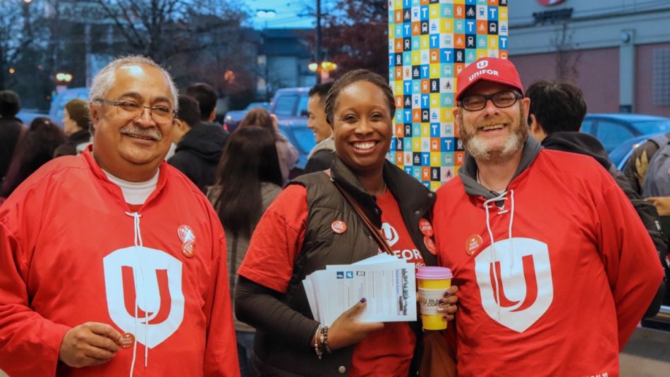 "Three Unifor members wearing Unifor swag smiling and standing outside at a busy bus stop."