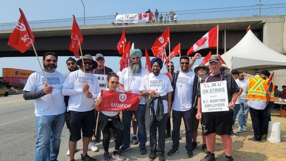 Many Unifor members posing outside with striking ILWU members. Unifor members are holding red flags and ILWU member is wearing an On Strike placard.