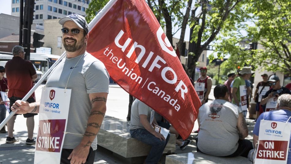 Man with sunglasses holding a Unifor flag and wearing an On Strike placard