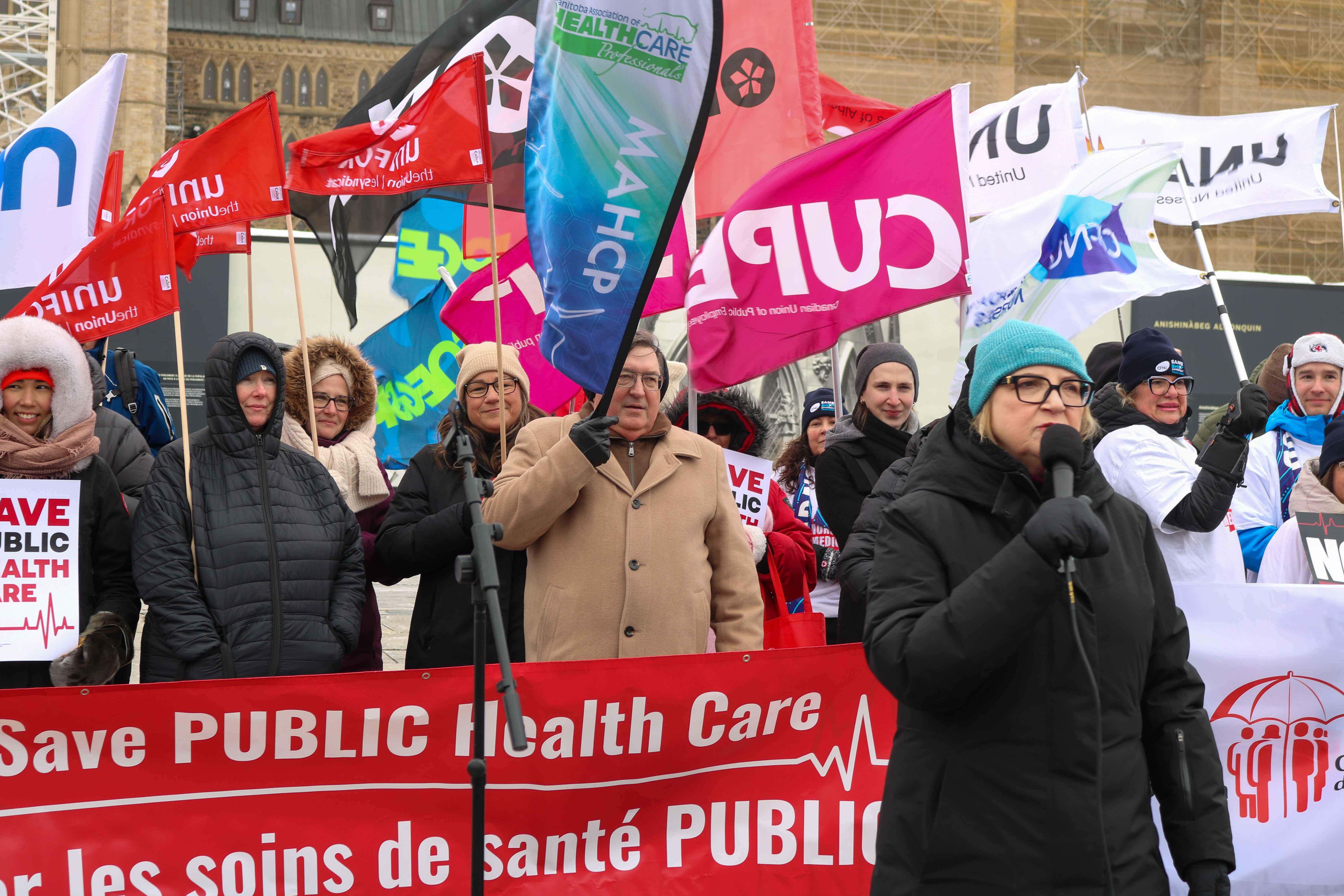 A women outside in winter gear speaking in front of a red banner and a large crowd