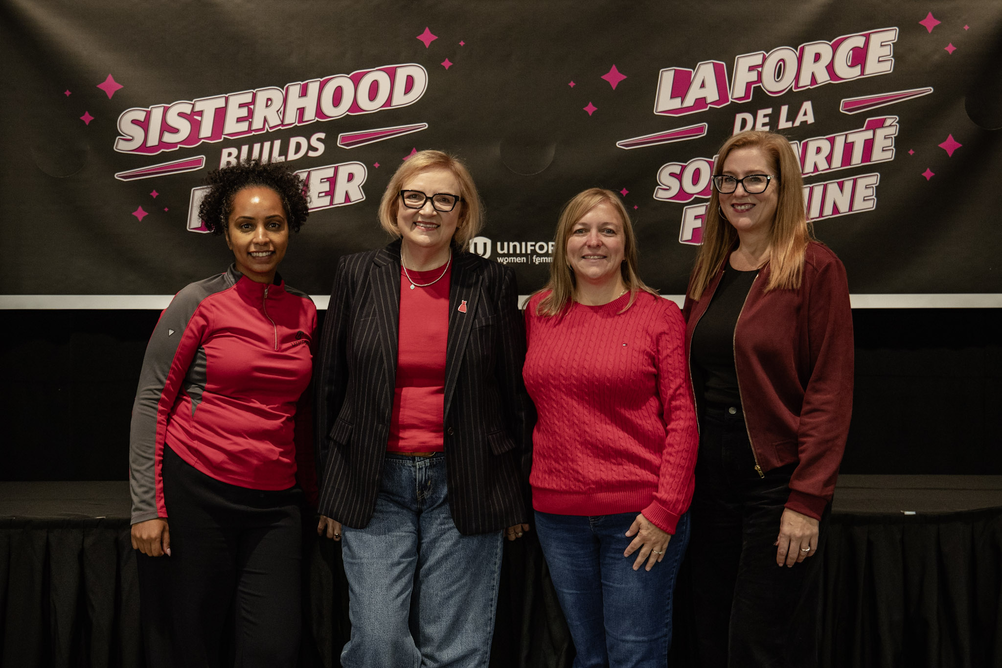 Four women pose in front of a banner that reads; sisterhood builds better, smiling 