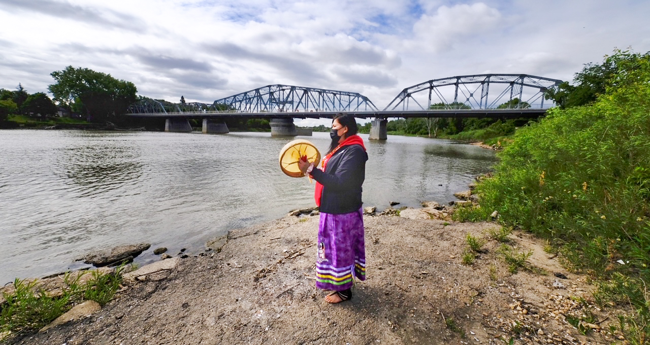 Woman standing on riverbank holding a drum. A bridge is visible in the background. Woman standing on riverbank holding a drum. A bridge is visible in the background.