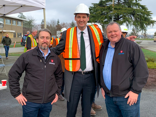 B.C. Premier David Eby posing for a photo in a high vis vest and a hard hat between Gavin McGarrigle and Travis Gregson.