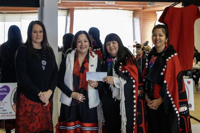 Four women, two wearing ribbon skirts and two wearing blankets with Indigenous styling pose holding a cheque.