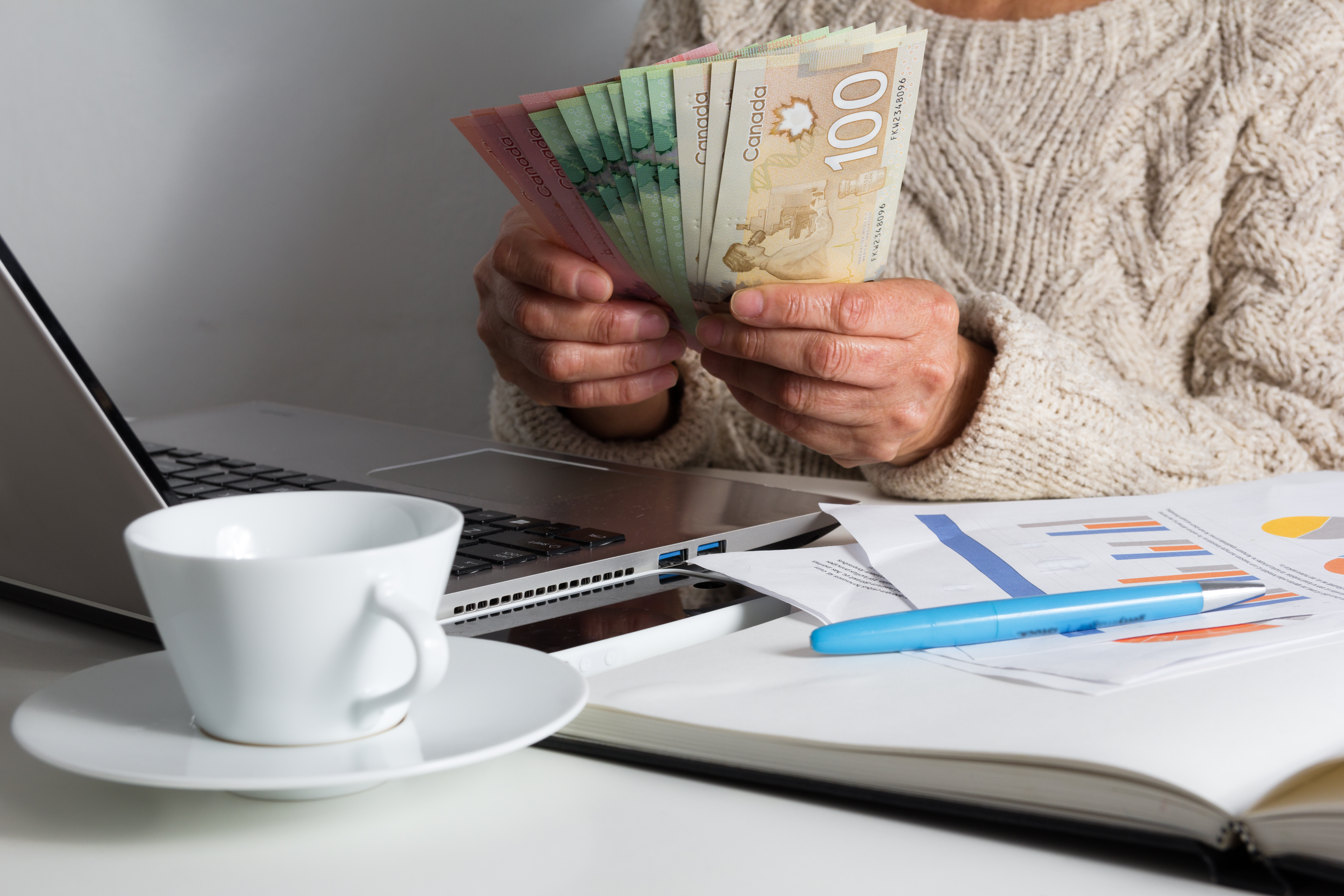 a person sitting at a laptop next to a notebook and a cup of tea holding money different bills fanned out