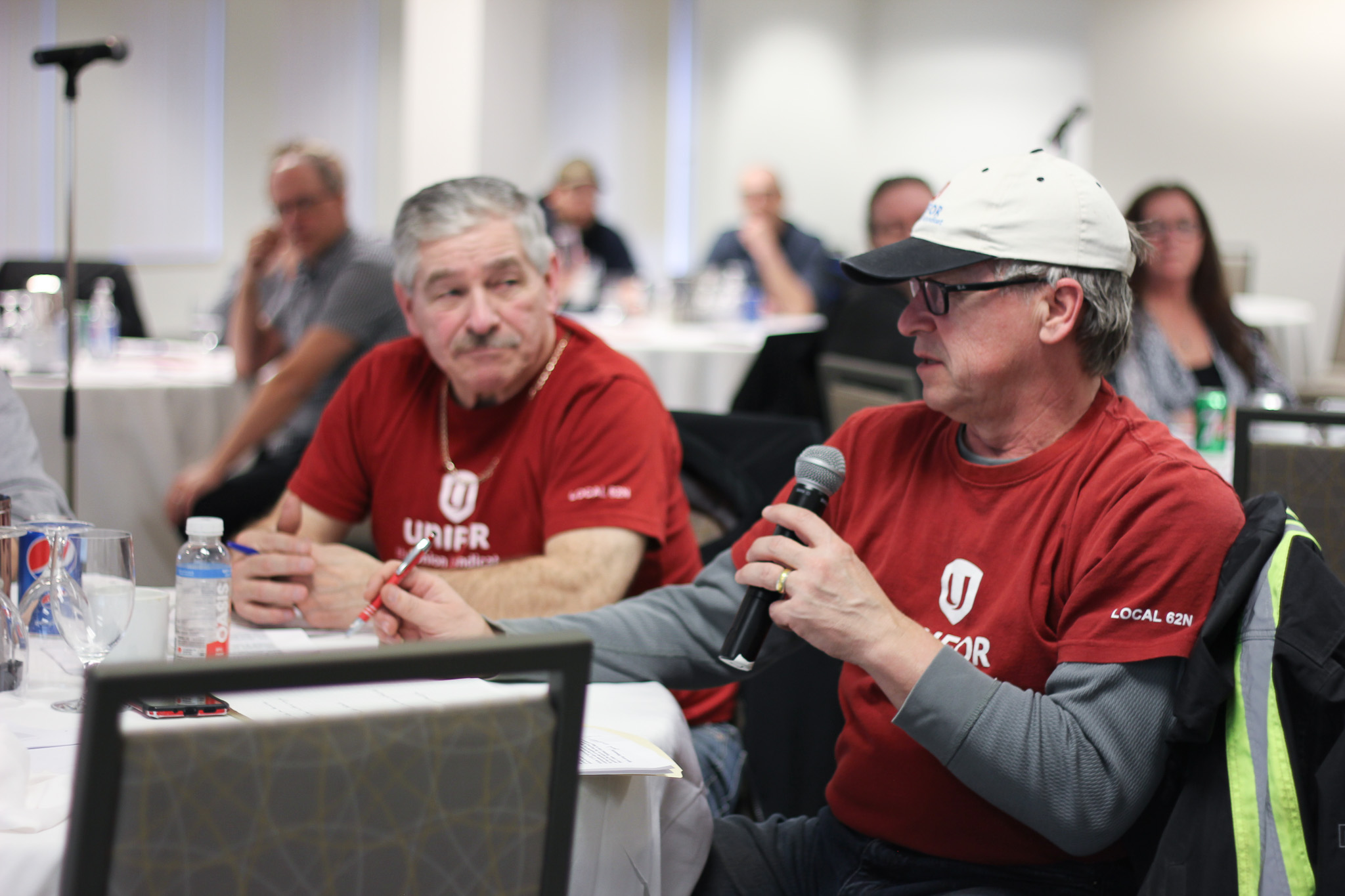 Two men sitting at a table wearing red Unifor tshirts, one is holding a mic and wearing a ballcap
