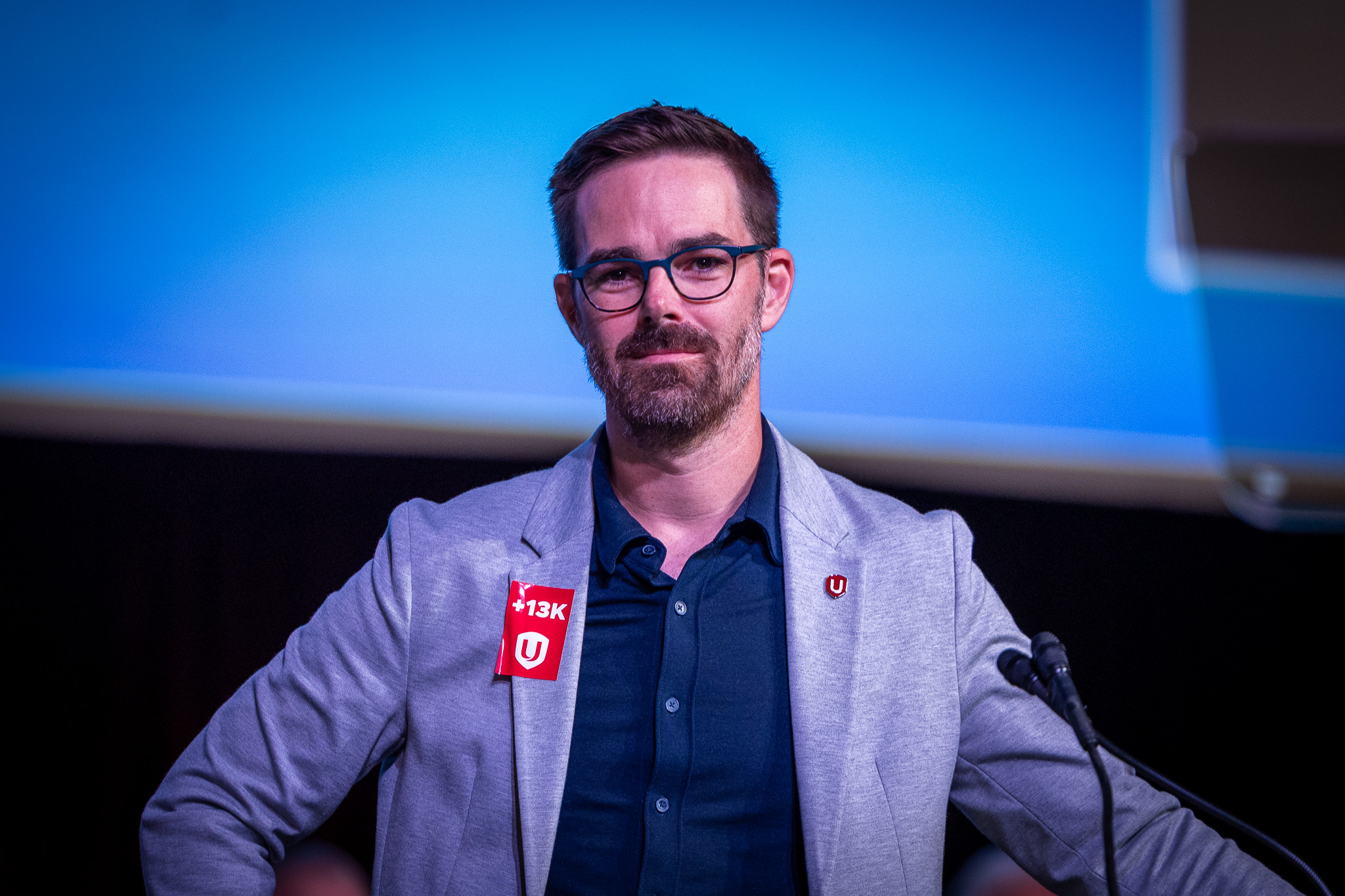 A man in a blue shirt and gray jacket, glasses digital screen in the background