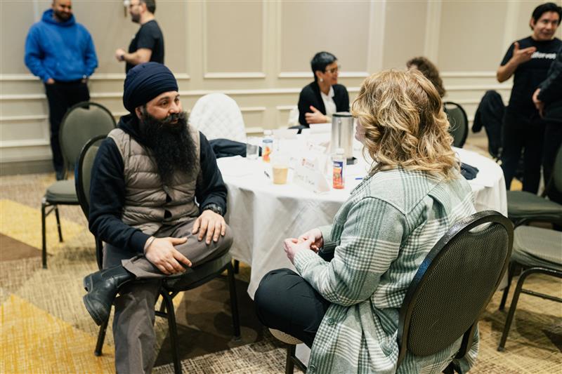 Two people sitting facing eachother beside a table with people in the background