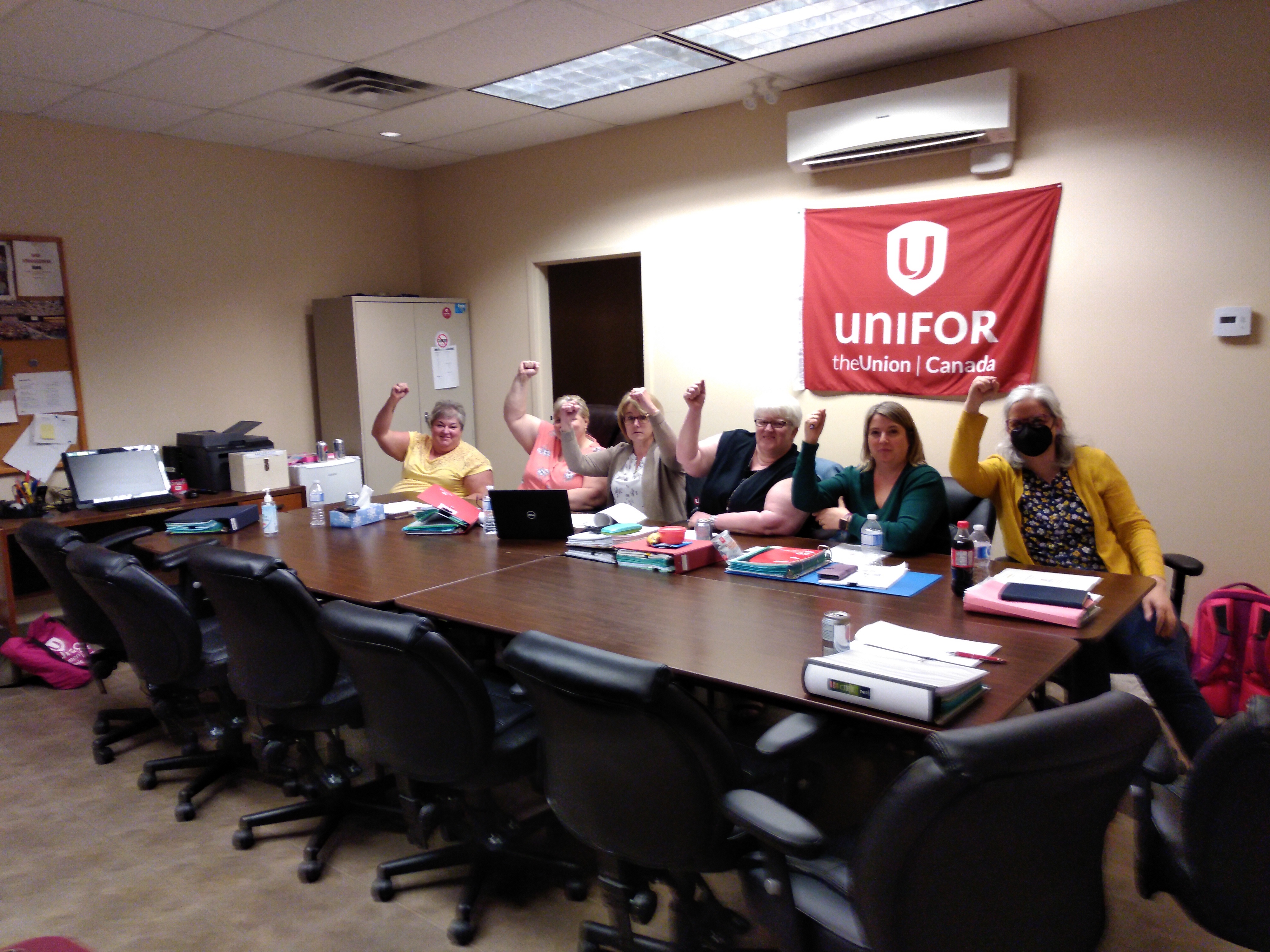 A group of women with their arms raised in the air, seated at a table. A red Unifor flag is shown on the wall behind.