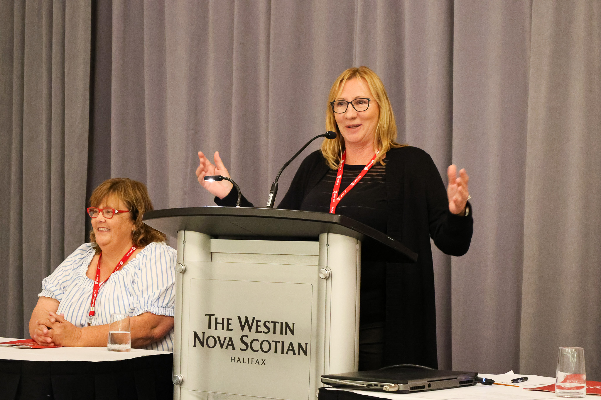 A women standing speaking at a podium, a women sits beside at a headtable