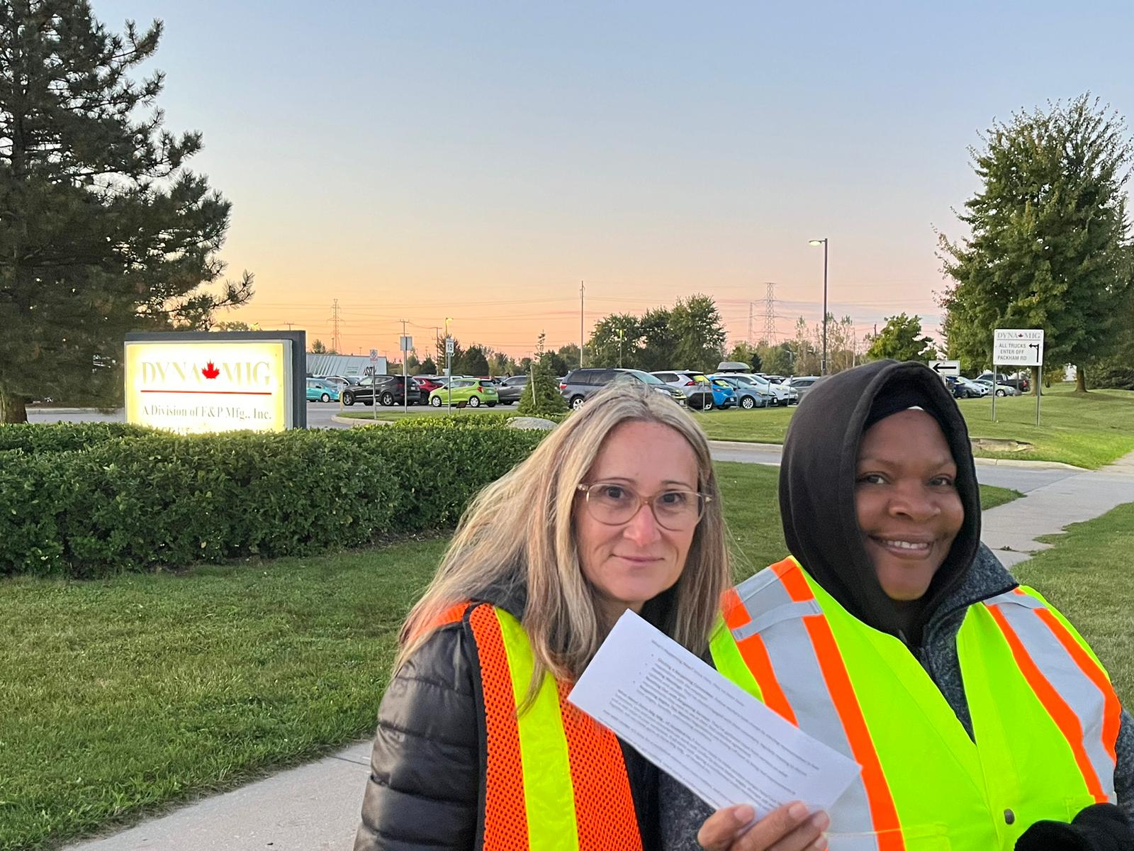 Two women in safety vests outside, a sign and lawn in the background