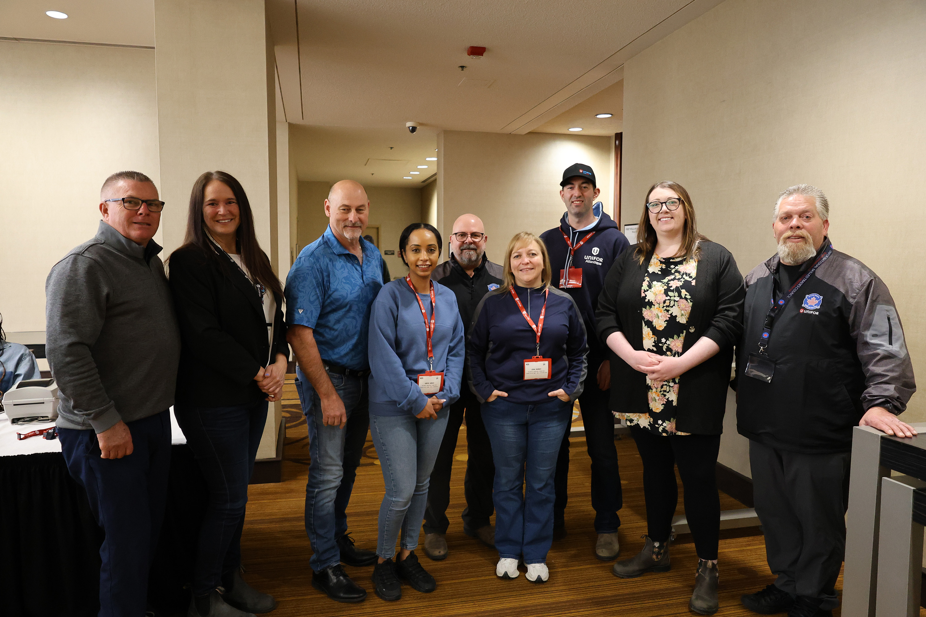 A group of nine people stand indoors, posing for a group photo at an event. Most are wearing conference name tags and casual attire.
