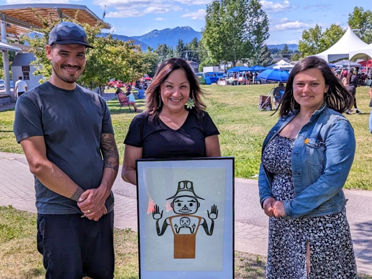 Nathan Woods poses with Unifor Staff Gina Smoke (L) and Nakkita Trimble (R) Three people standing outside with framed artwork.