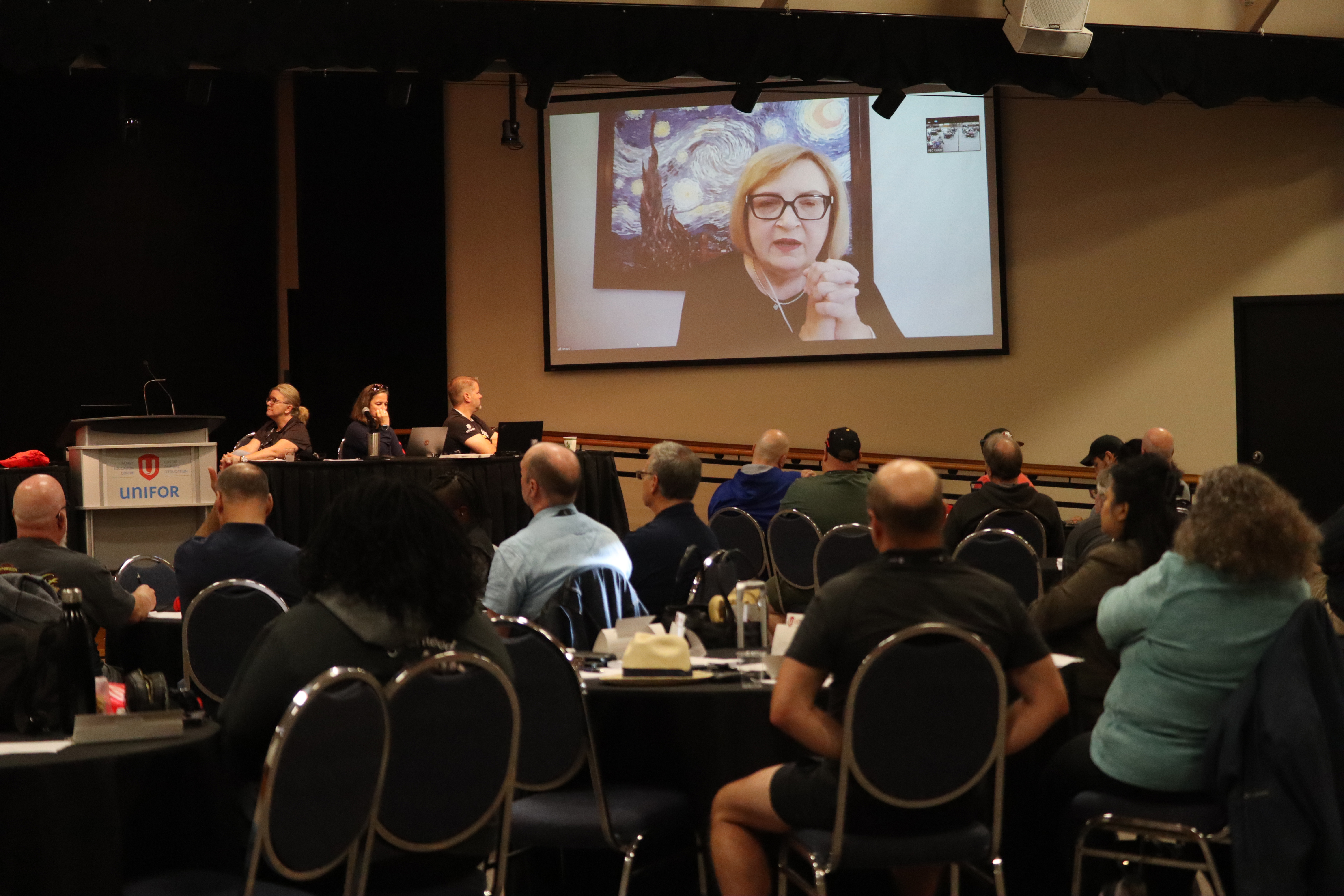 a dark room full of people sitting in chairs looking at a women on a large screen