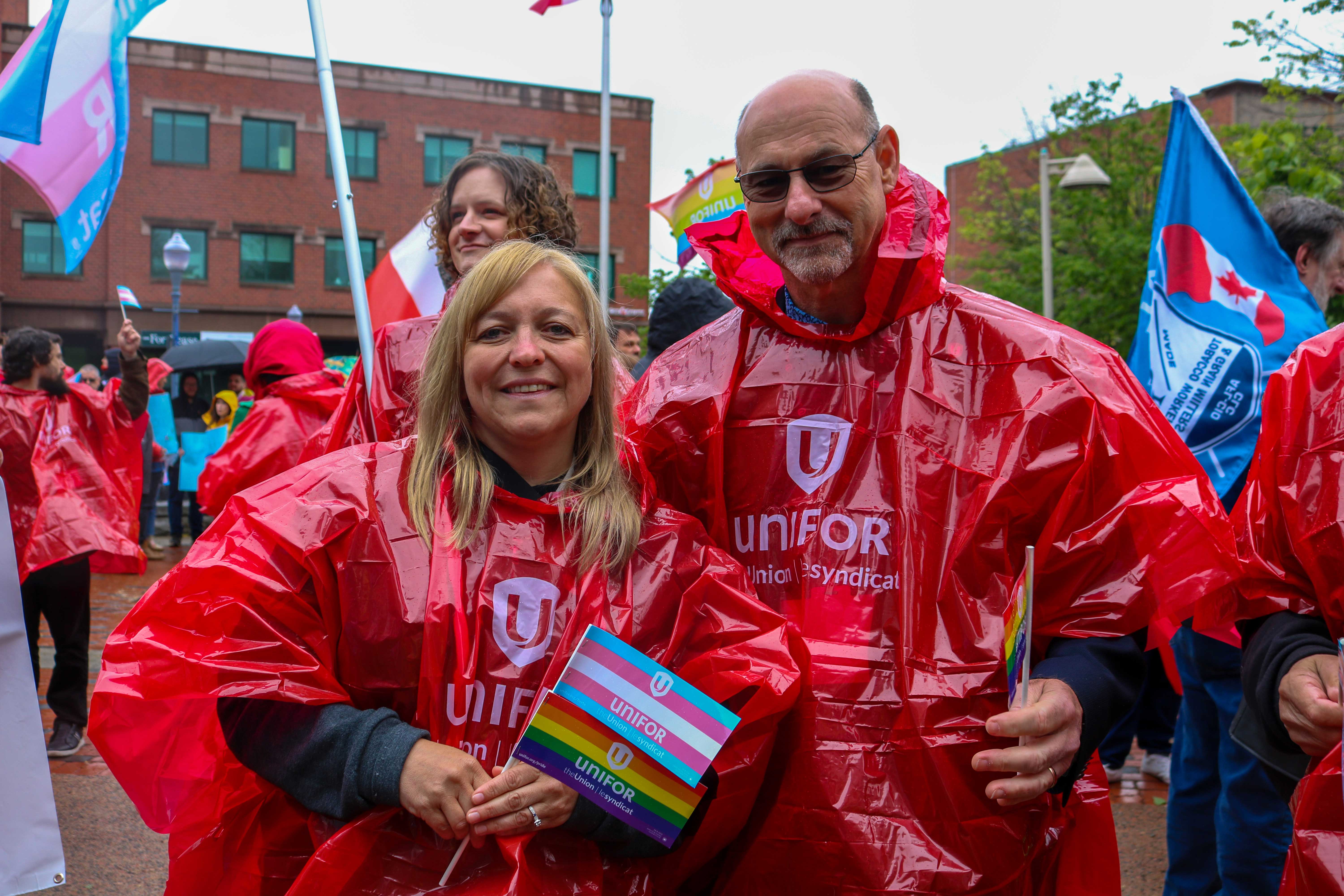 Jen and Len holding a trans pride flag wearing rain ponchos