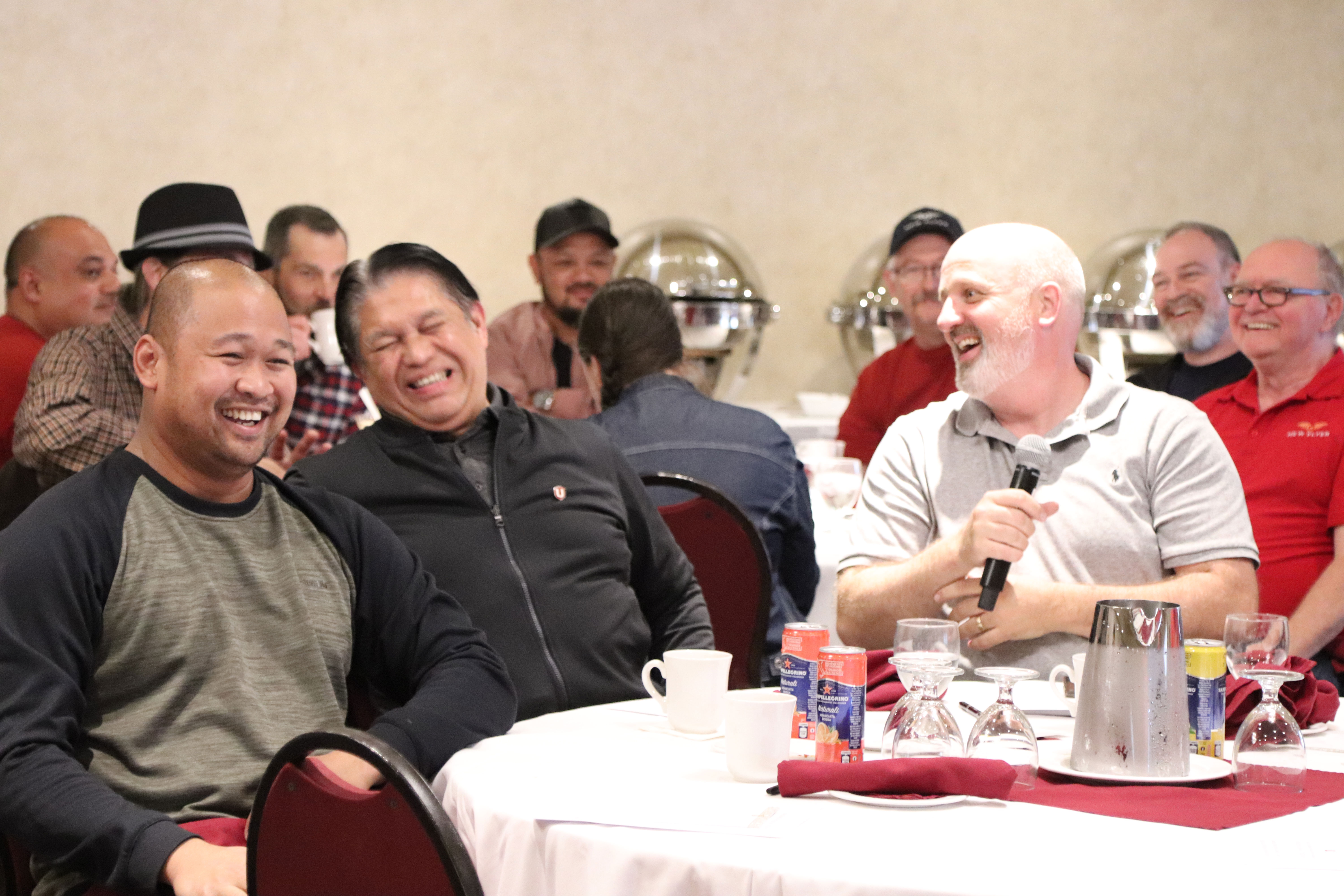 three men sitting at a table laughing one is taking into a mic