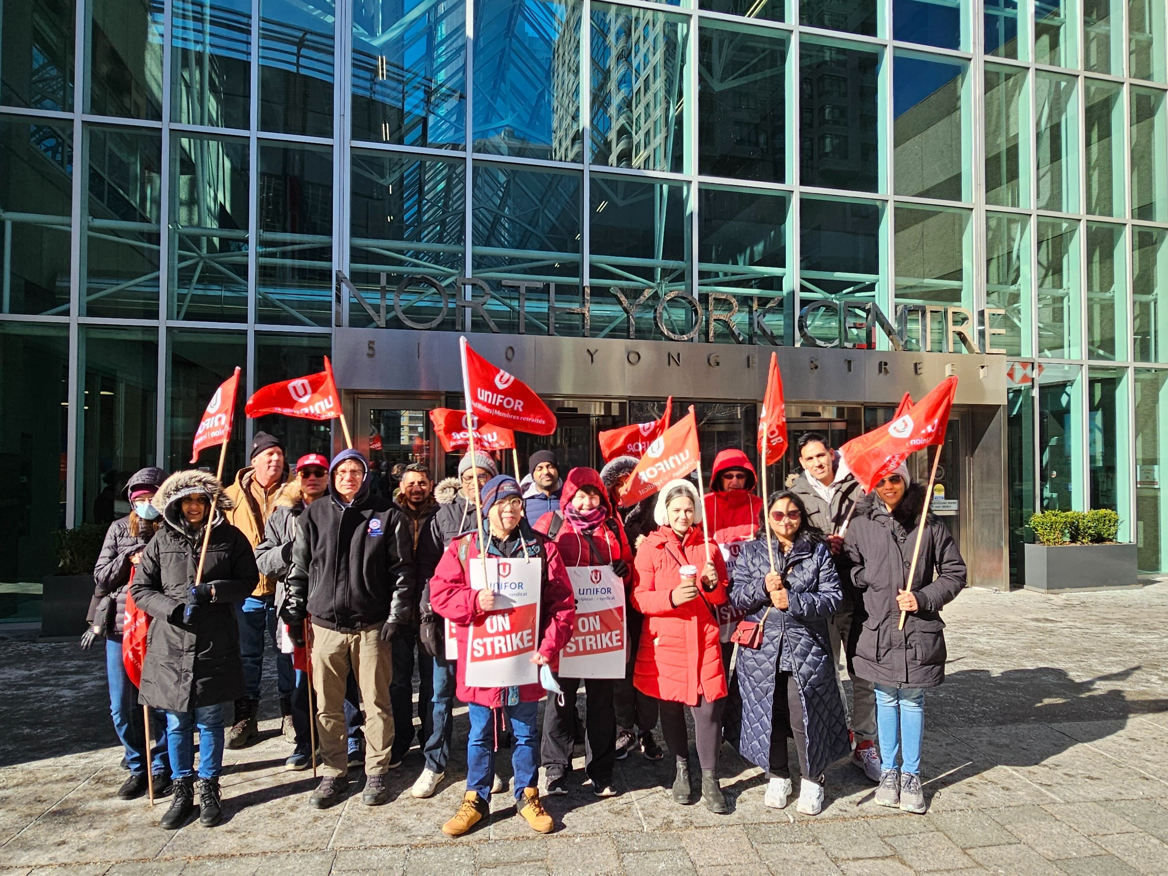A large group of people standing in the sun wearing winter cloths holding picket signs and Unifor flags.
