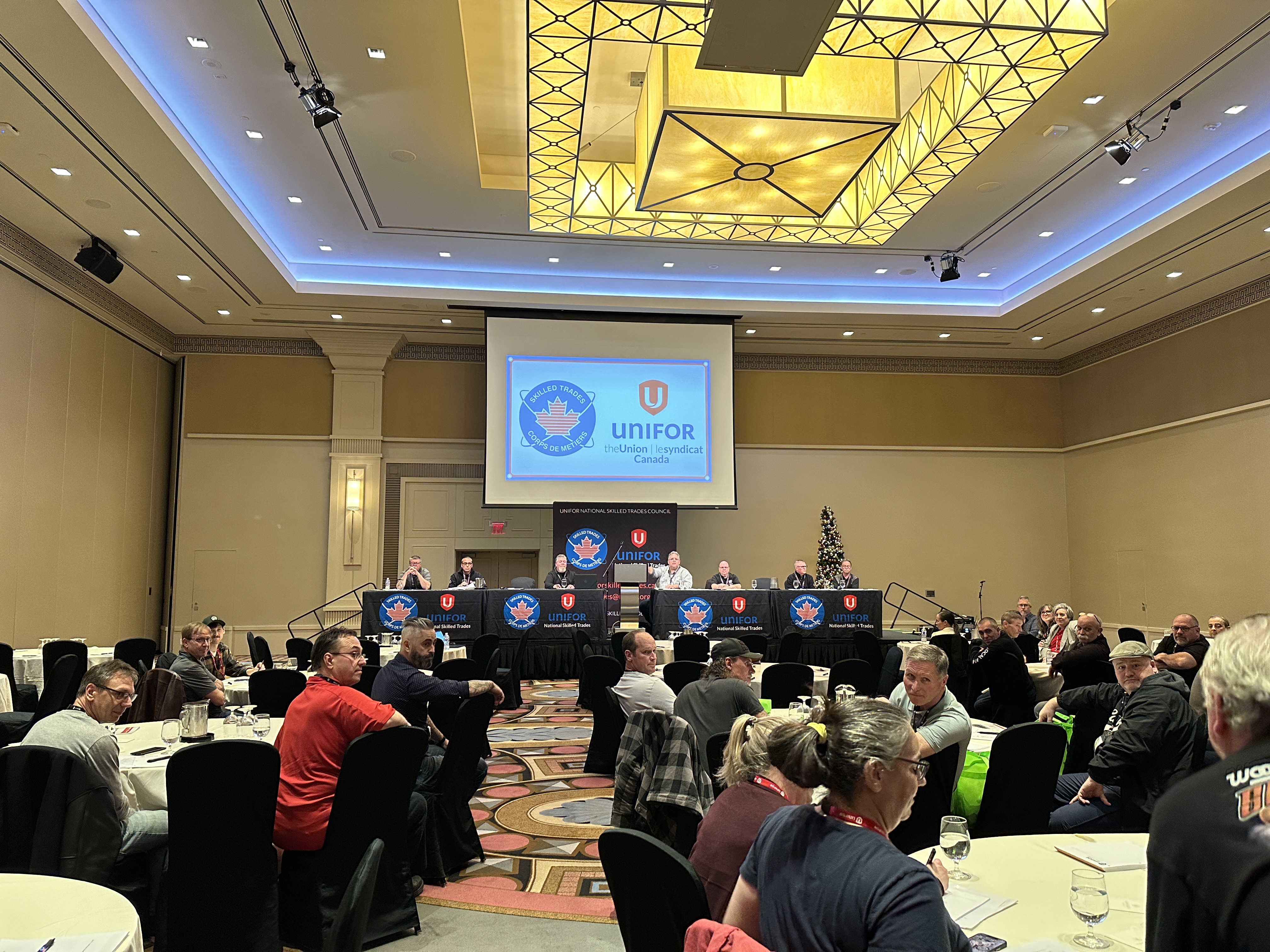 people sitting at tables looking front at a screen with a Unifor Skilled Trades Logo a red flag with a blue circle around it.