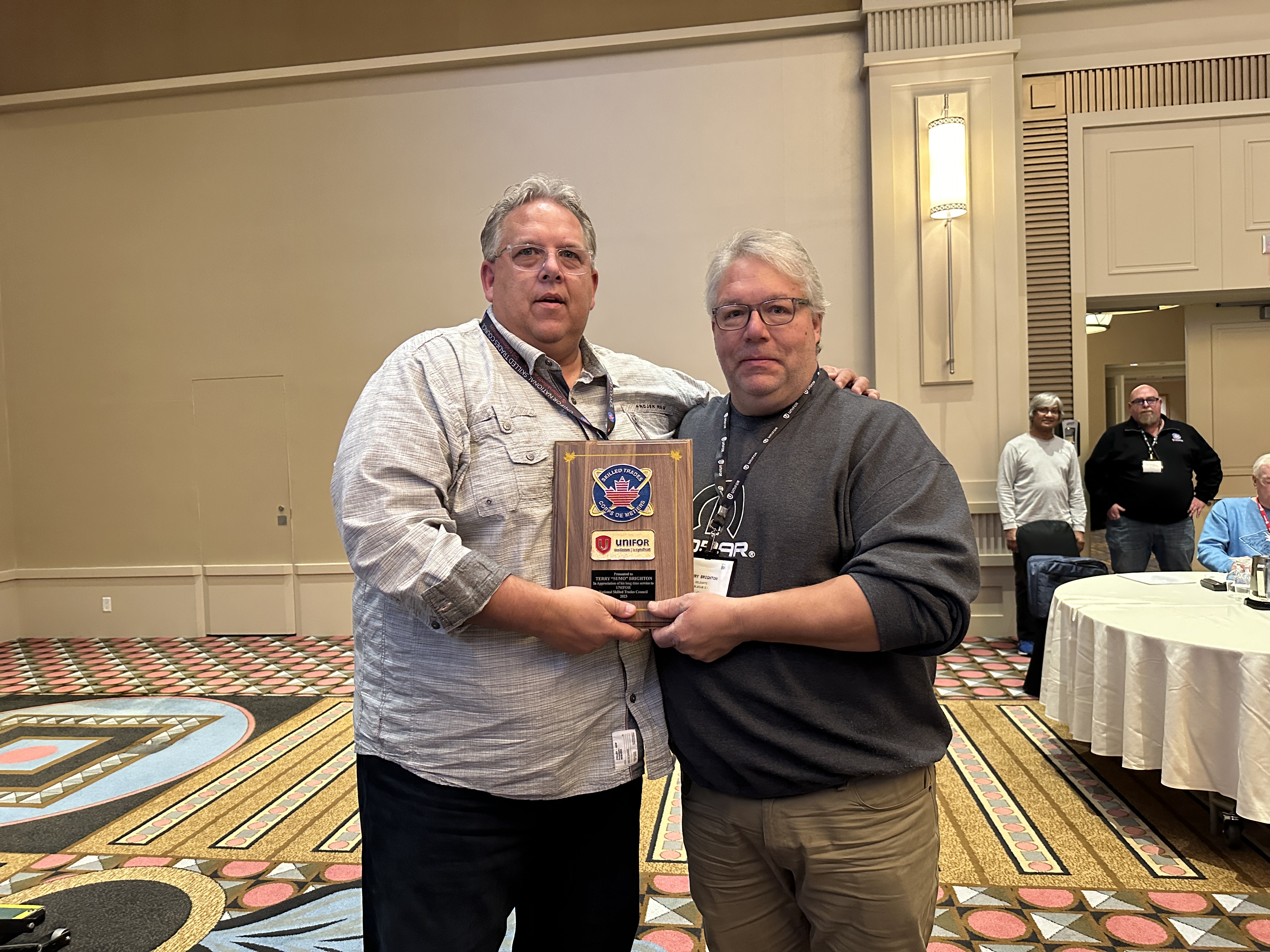 Two man standing arms around each other holding a plaque