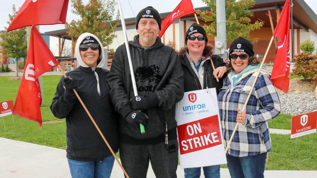 Four people posing for a photo outside with Unifor flags and an On Strike placard