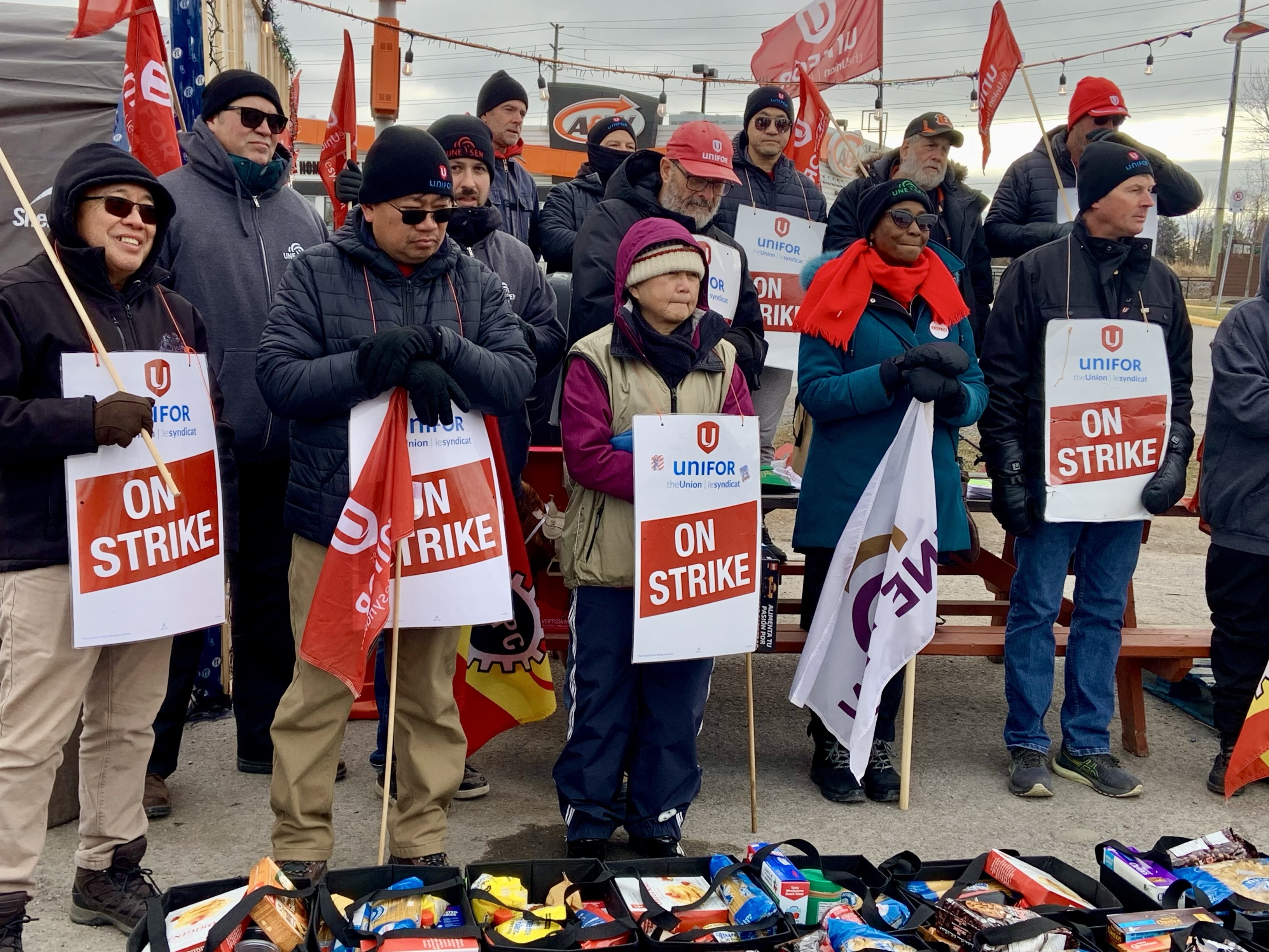 A group of people holding Unifor flags and 'On Strike" signs stands behind food hampers on the ground.