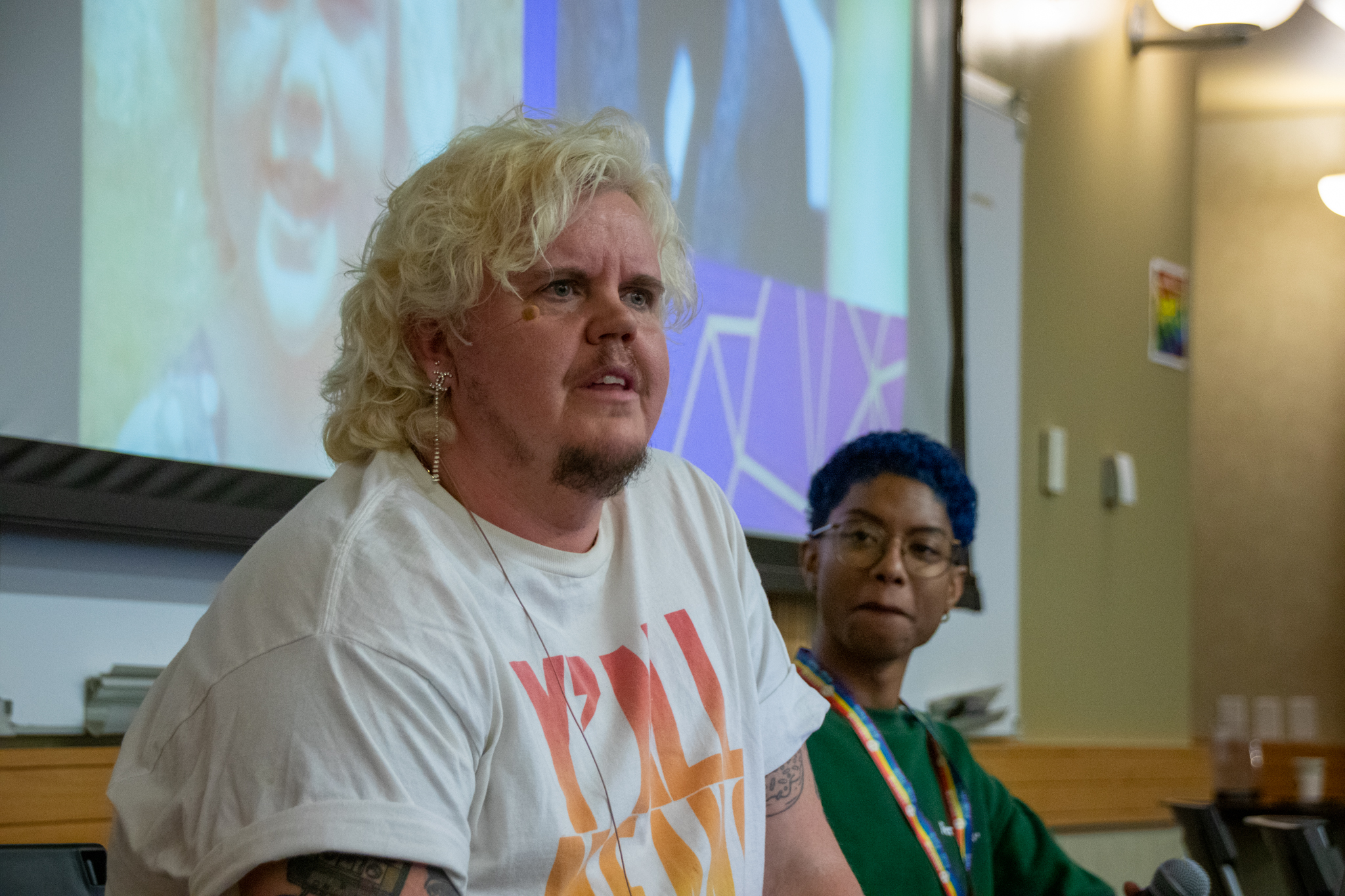 Kaleb Robertson, who has blonde hair and is wearing a white t-shirt, speaks, while a person behind them looks. In the background is a projection screen with Kaleb’s photo.