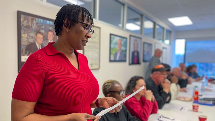 Person standing and reading off of a piece of paper with others listening seated in the background.