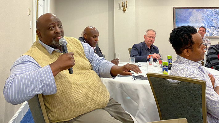 Man seated speaking into a microphone. Others seated at round table nearby.