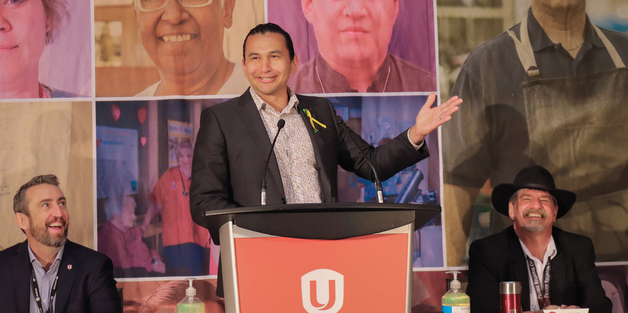 Wab Kinew smiling at a podium with Gavin McGarrigle and Guy Desforges seated on either side.