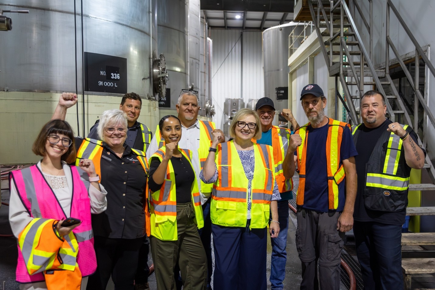 Nine people with fists raised standing in a wine crushing producation area of a winery