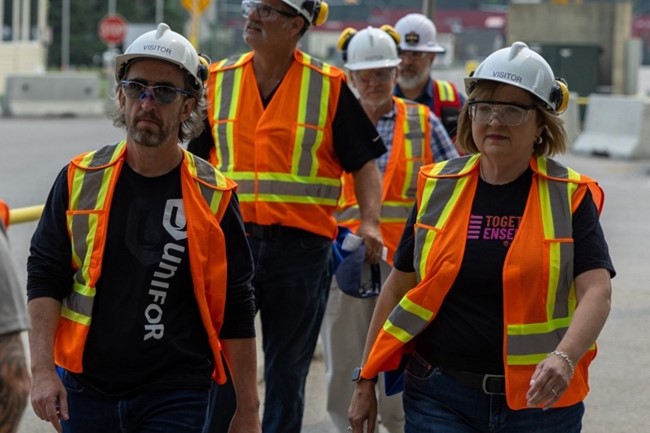 Des dirigeants d'Unifor portant des gilets de sécurité orange, des lunettes de sécurité et des casques de protection marchent à l'extérieur.