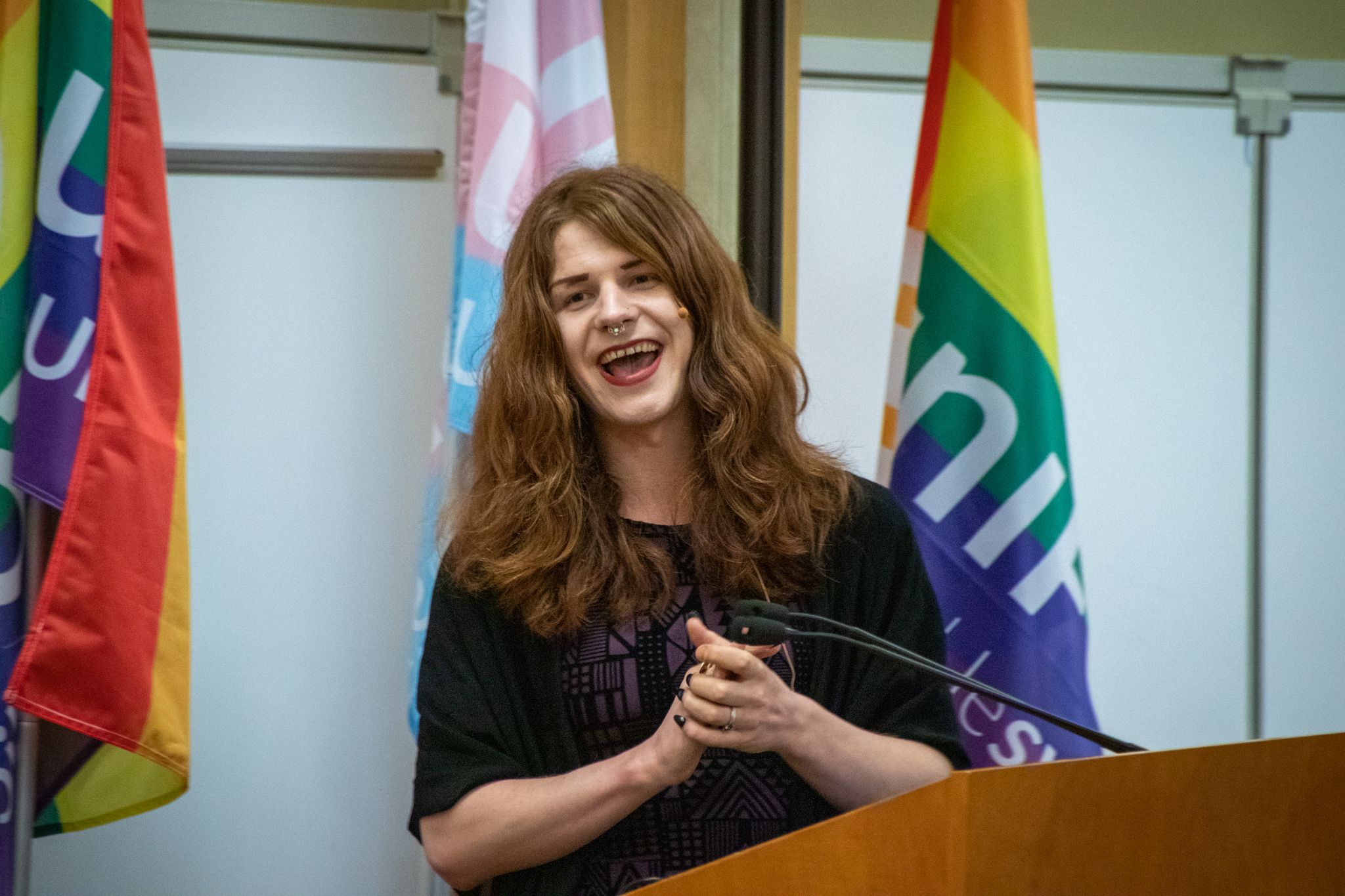 Fae Johnstone, who has long brown hair, a septum piercing and is wearing black nail polish, laughs while standing at a podium with Unifor rainbow flags in the background.