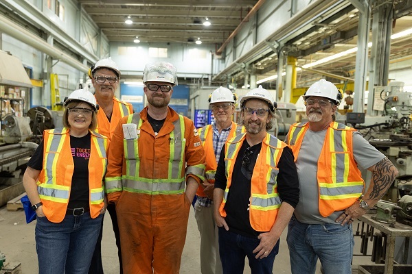 •	A group of people wearing safety vests and helmets stand in front of machinery at pulp and paper mill.