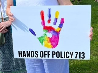 A person holding a sign of a colourful hand print that reads, Hands off policy 713