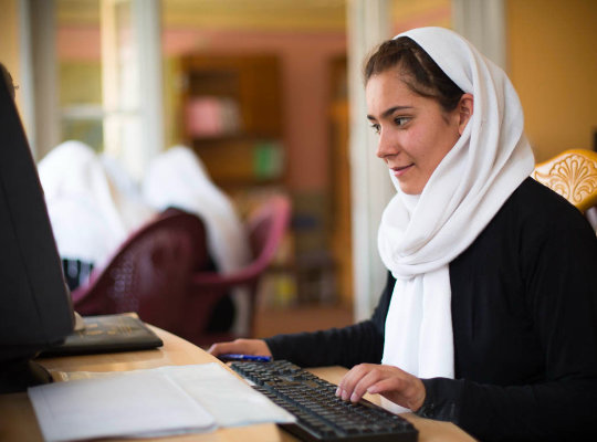 Young Afghan woman in a white headscarf works at a computer in a classroom, with other students seated in the background.	