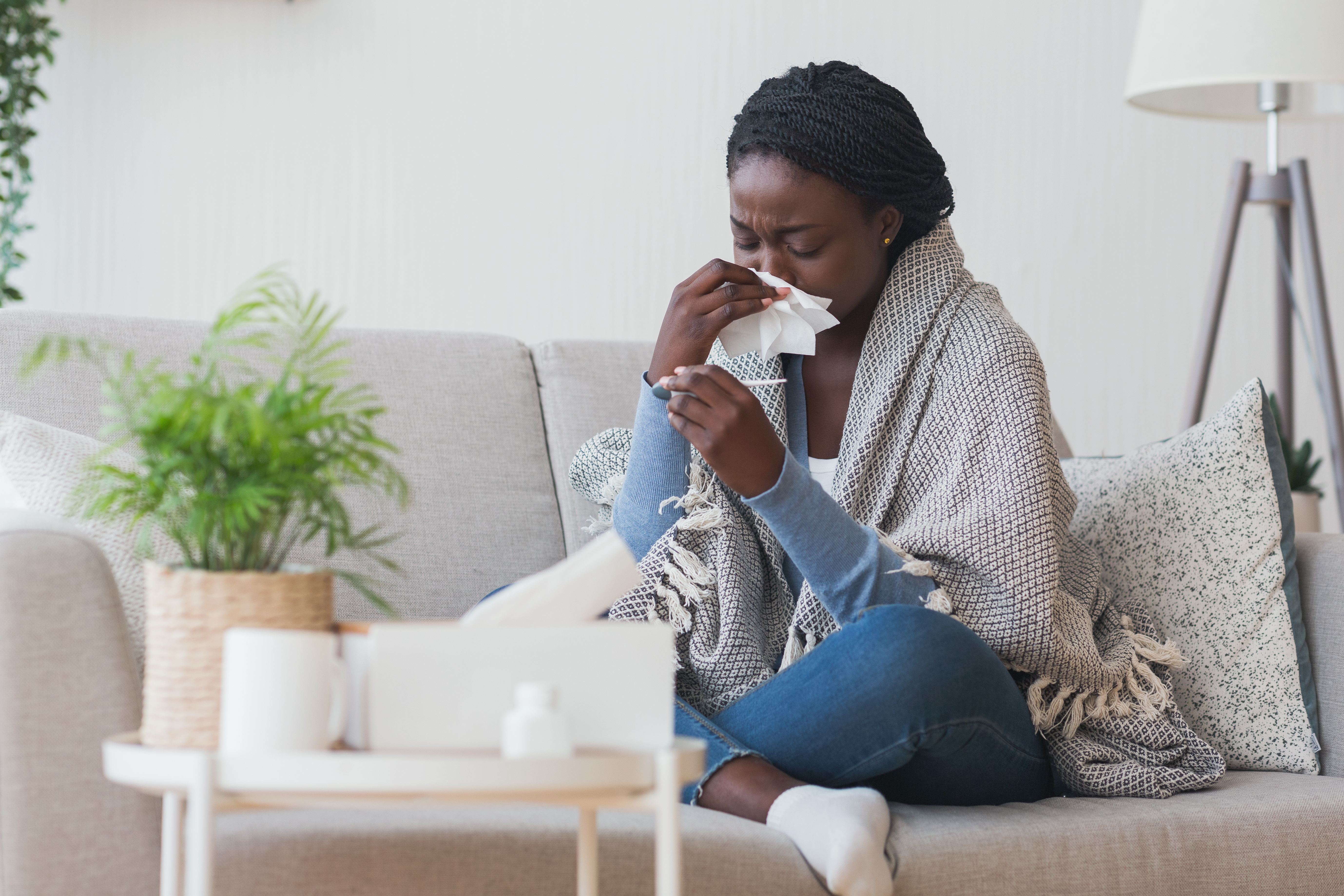 Woman on couch wiping nose with tissue and looking at thermometer.
