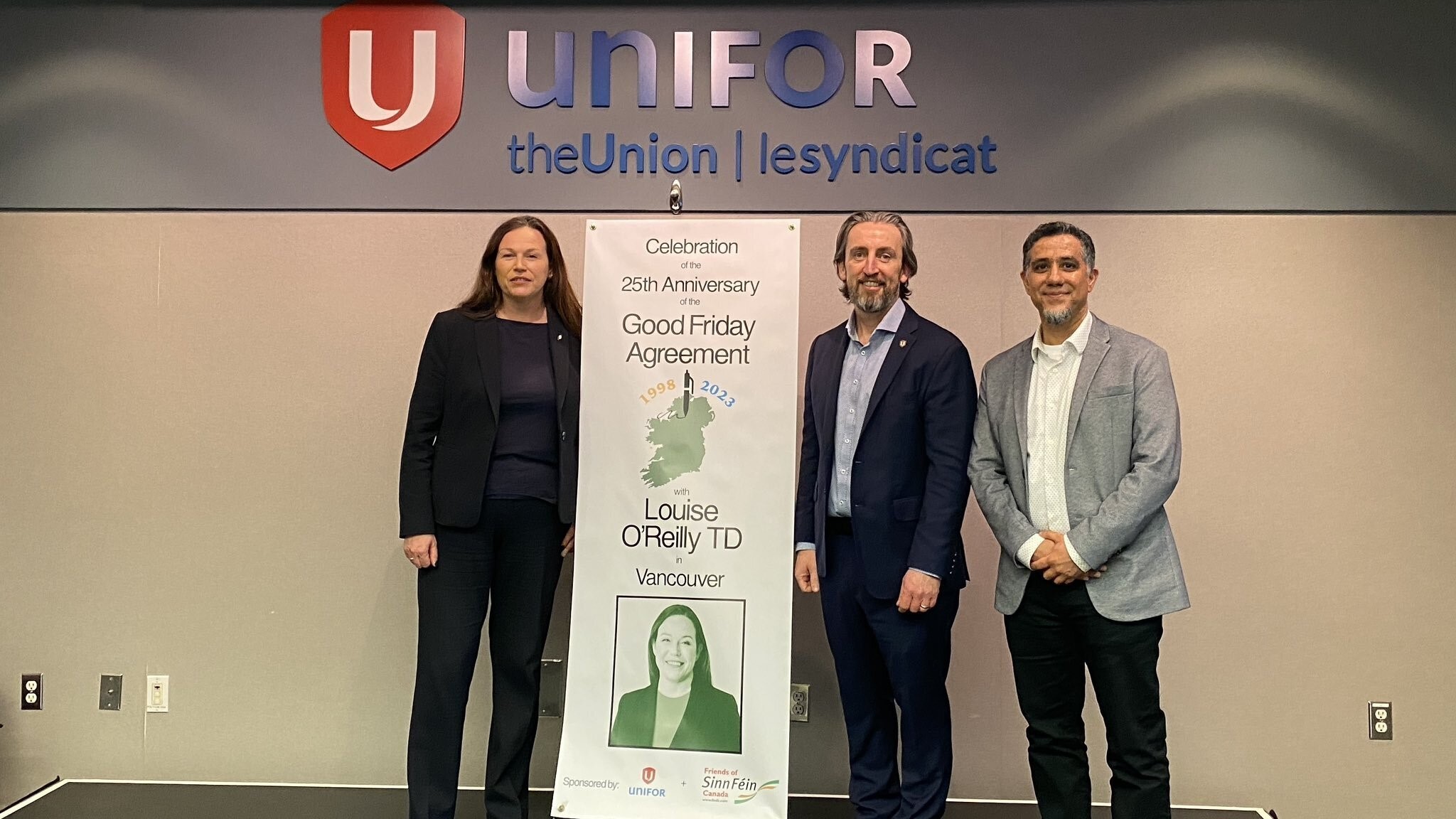 Two men and a woman stand in front of a sign that says, “Good Friday Agreement.” A Unifor sign is above the three of them.