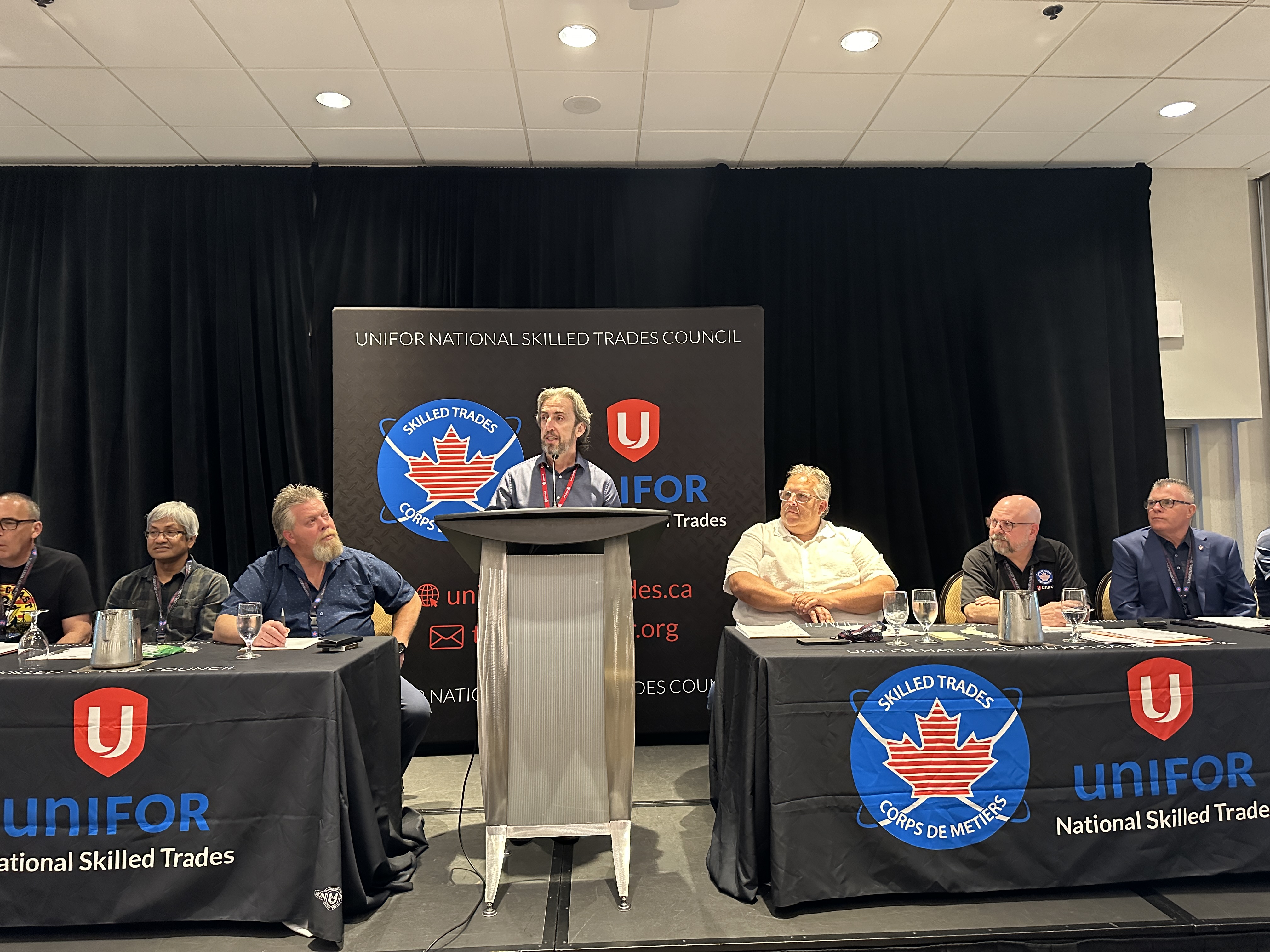 Man standing at a podium, three people sit at tables on either side