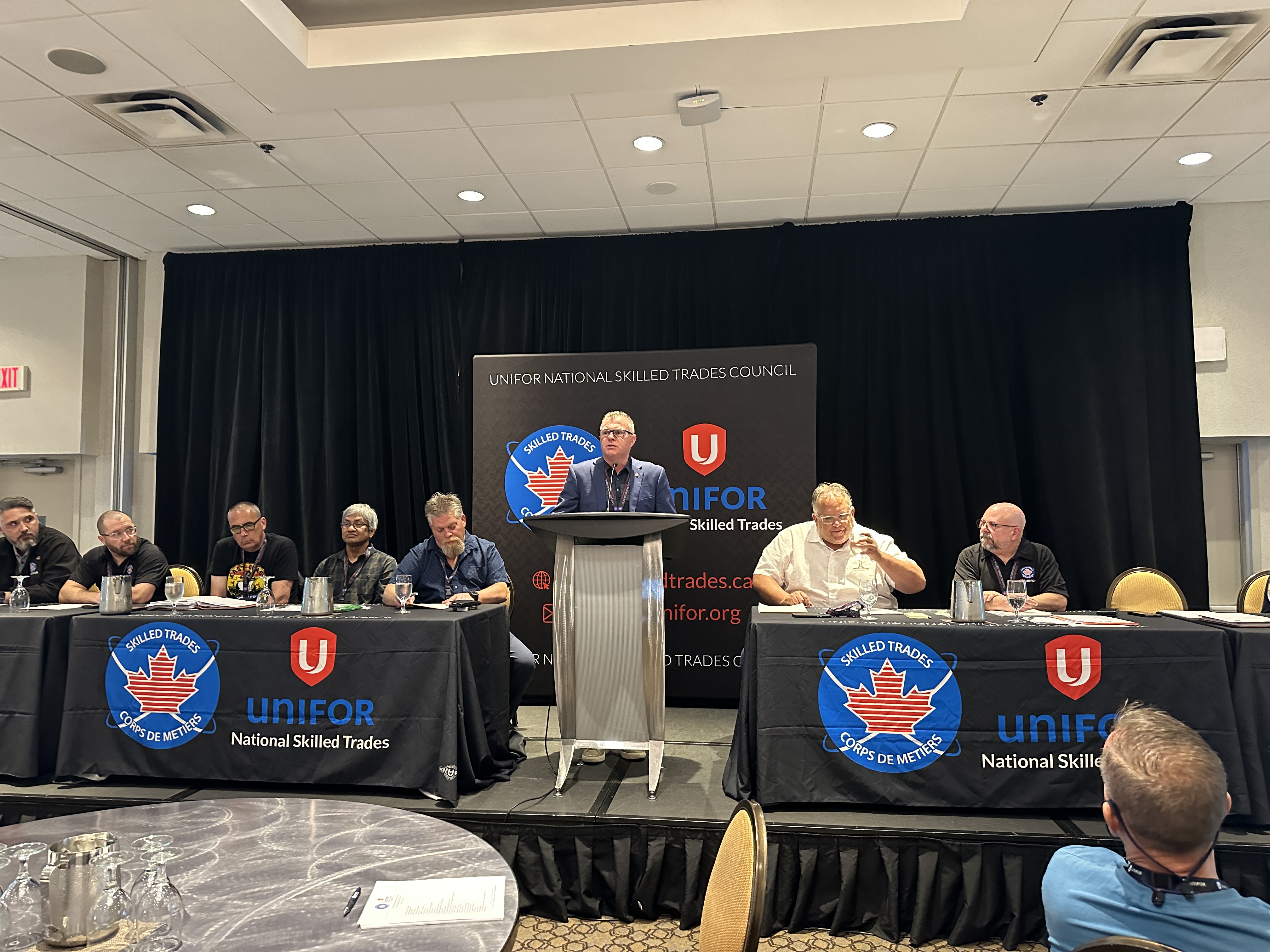 Man stands at a podium, three people sit at tables on either side