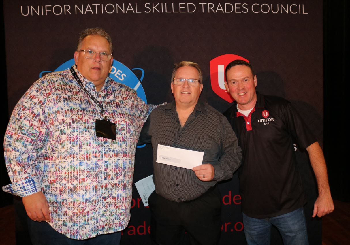 Three men standing together. Man in middle is holding a cheque