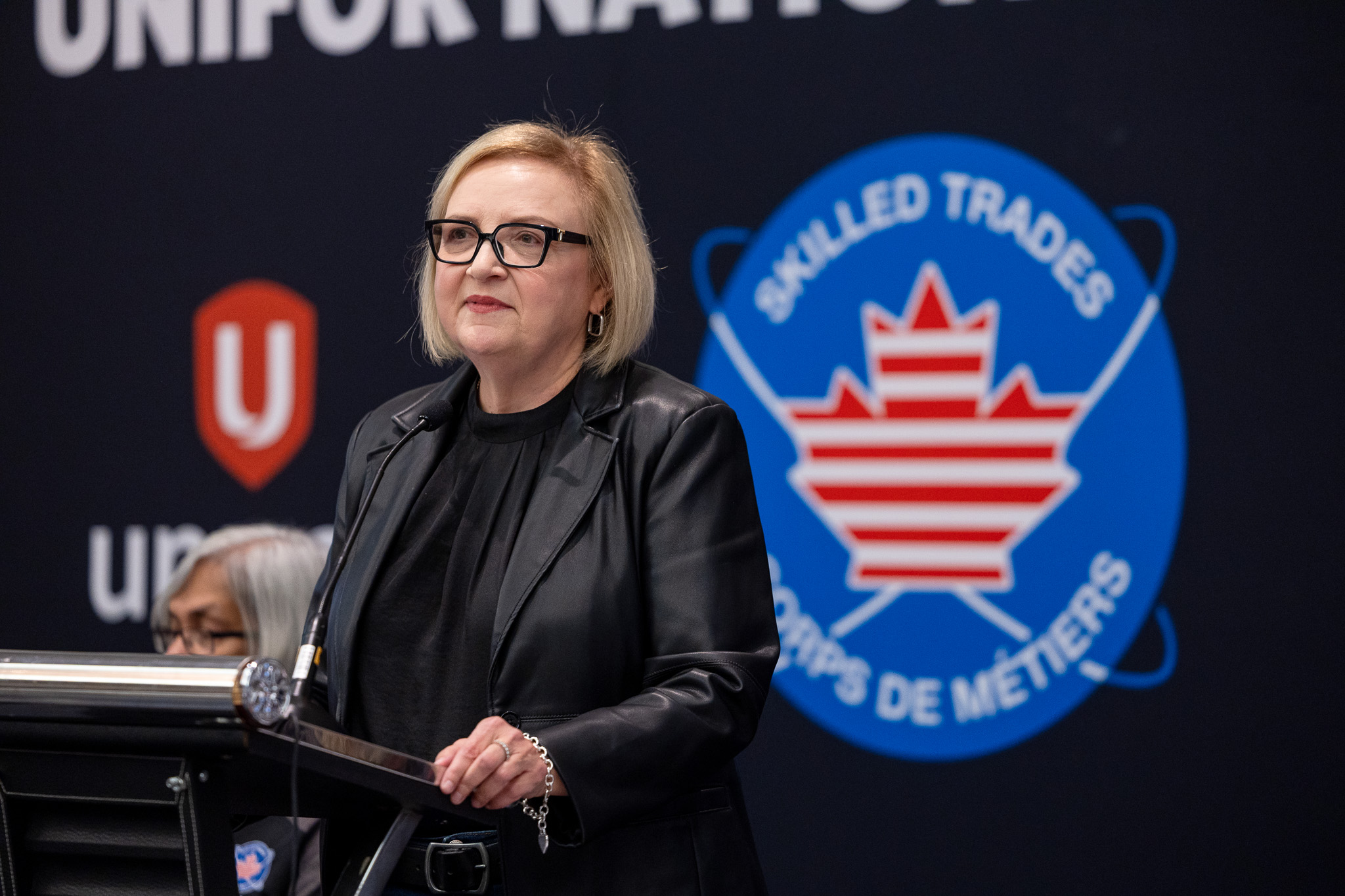 A women speaking at a podium the Unifor logo and skilled trades logo on a black background