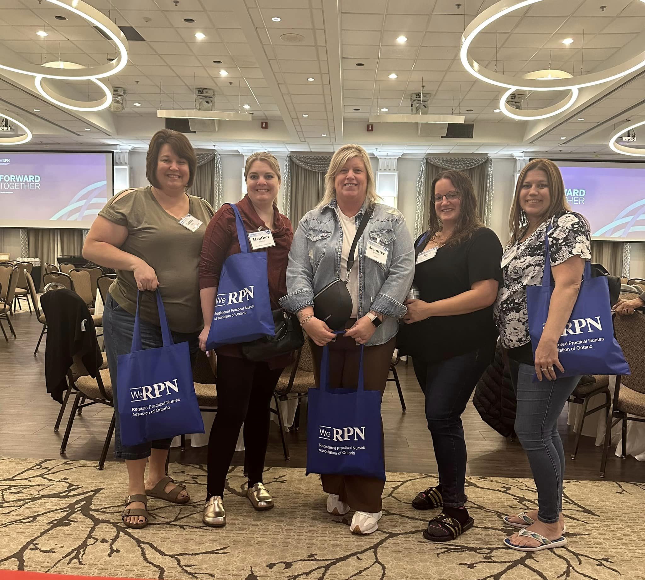 five women posing in a lobby with tote bags