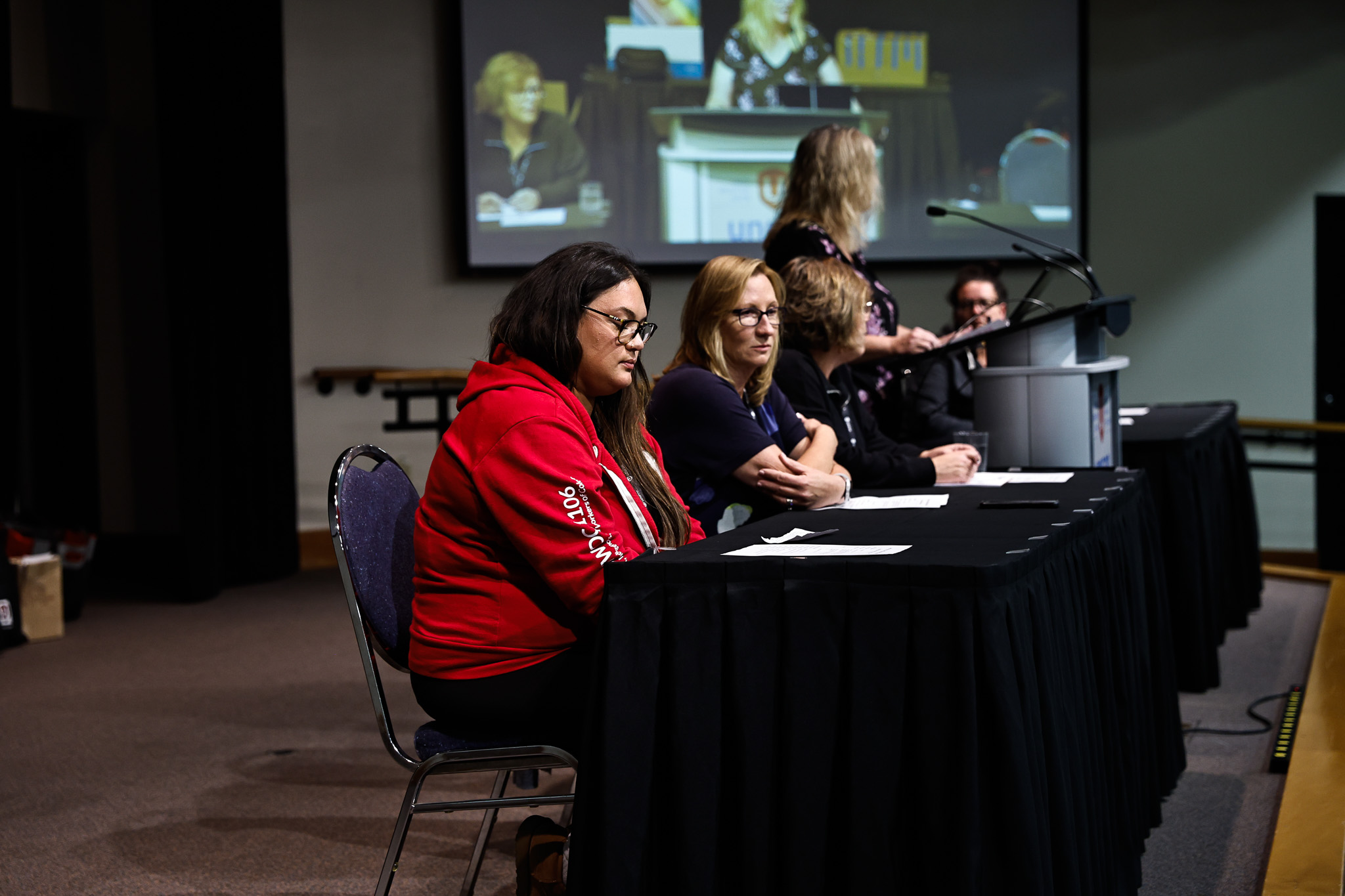 A head table with three poeple sitting, one women standing, speaks at the podium