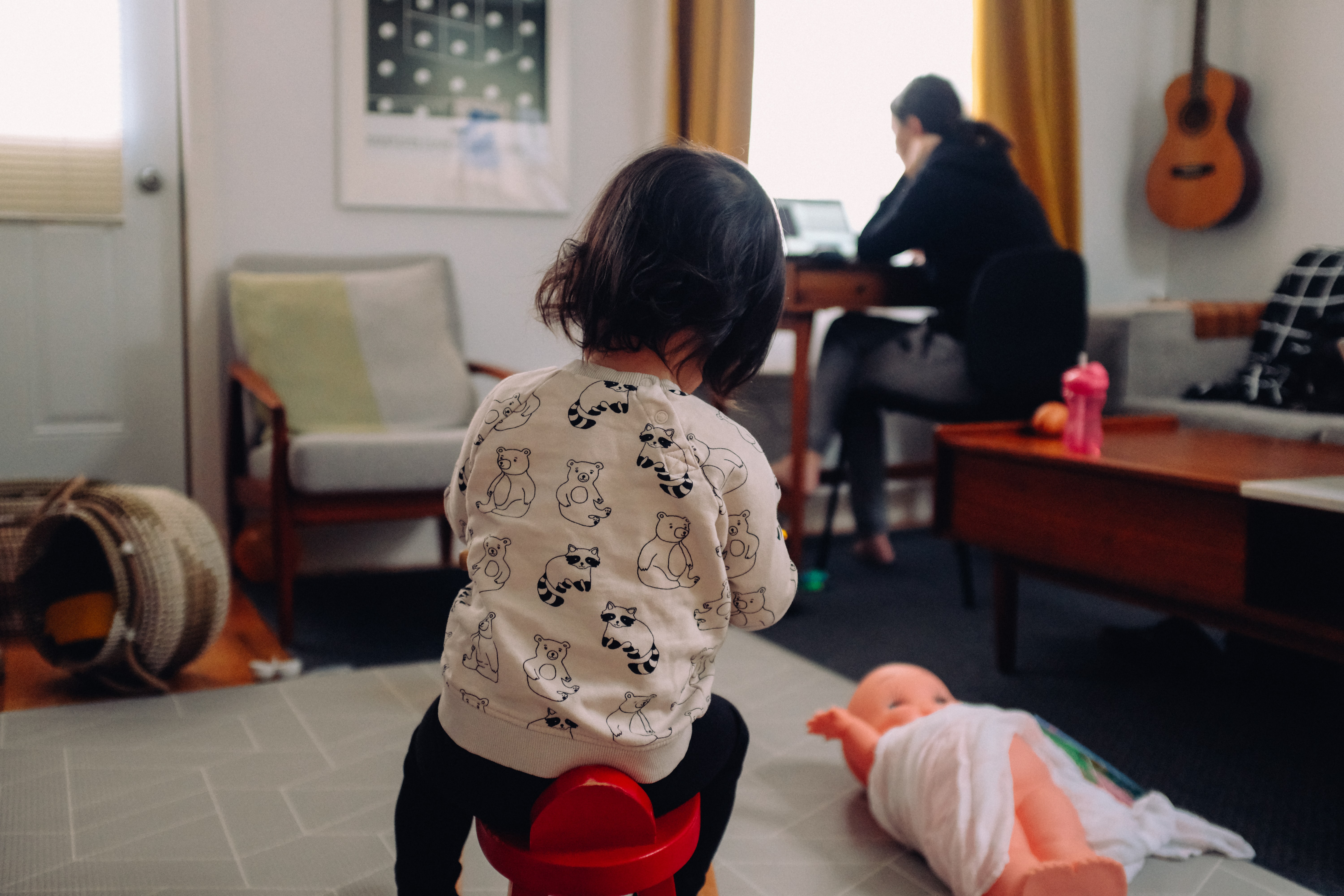 a child sitting on a toy with a parent on a computer in the background