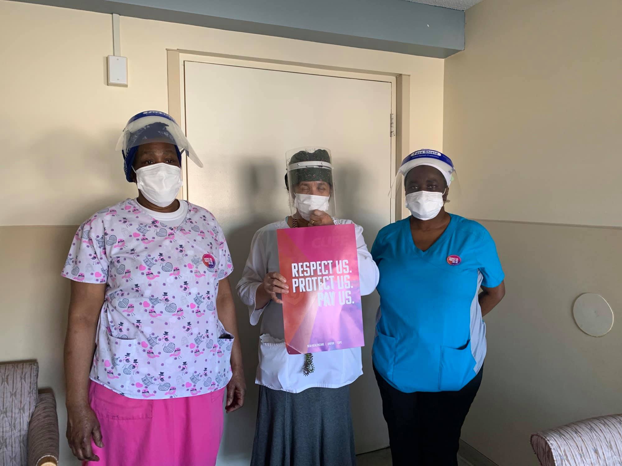 three diverse health care workers wearing masks and faceshields the women in the middle is holding a respect us, protect us, pay us sign