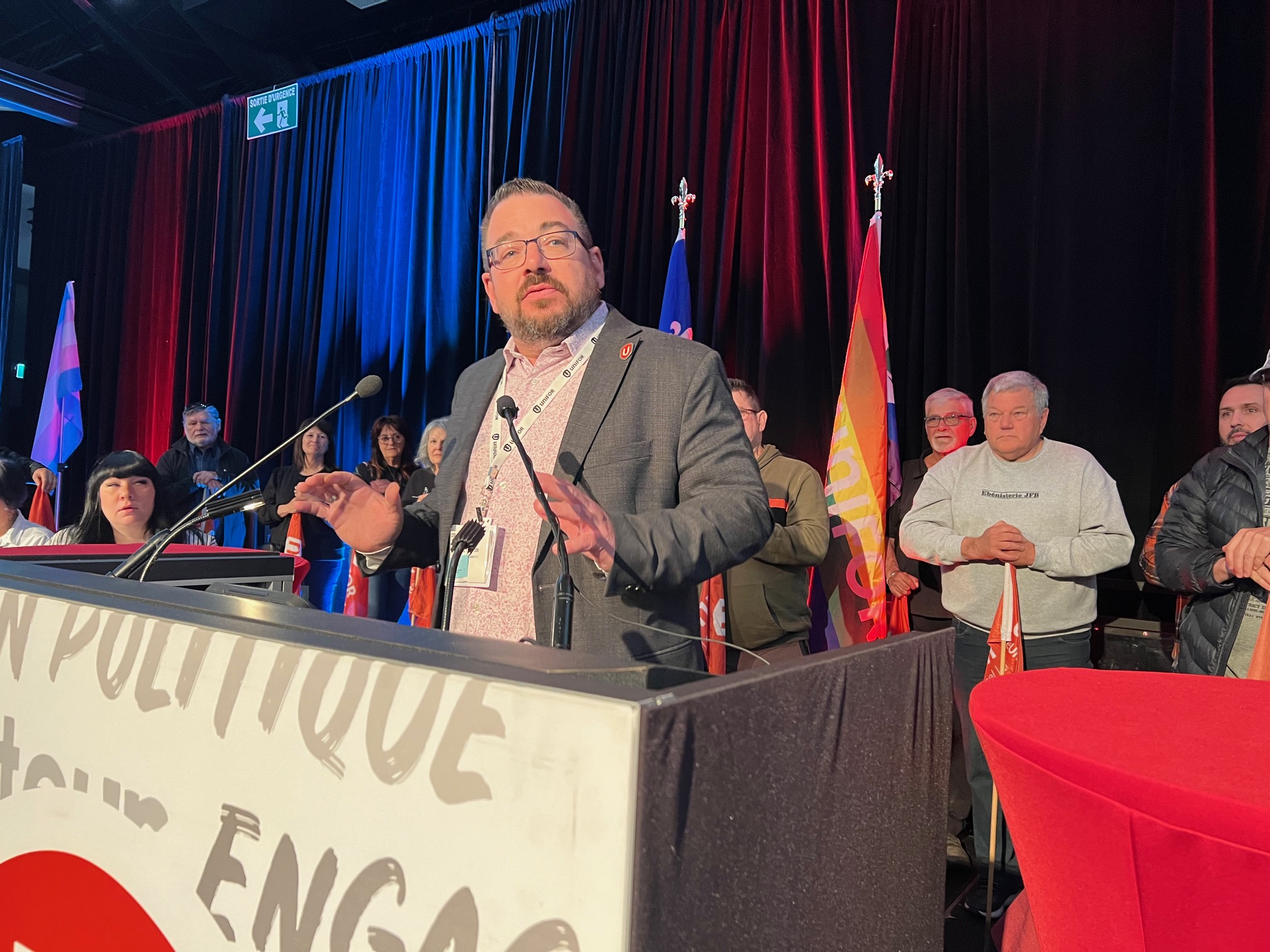 A man wearing glasses and a grey blazer speaks at the podium, while people, who are seated, watch in the background.