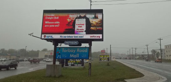 A billboard shows an image of a Local 597 member wearing a sign that reads "Locked out 79 weeks in Gander" next to the text "Premier Dwight Ball, whose side are you on?"