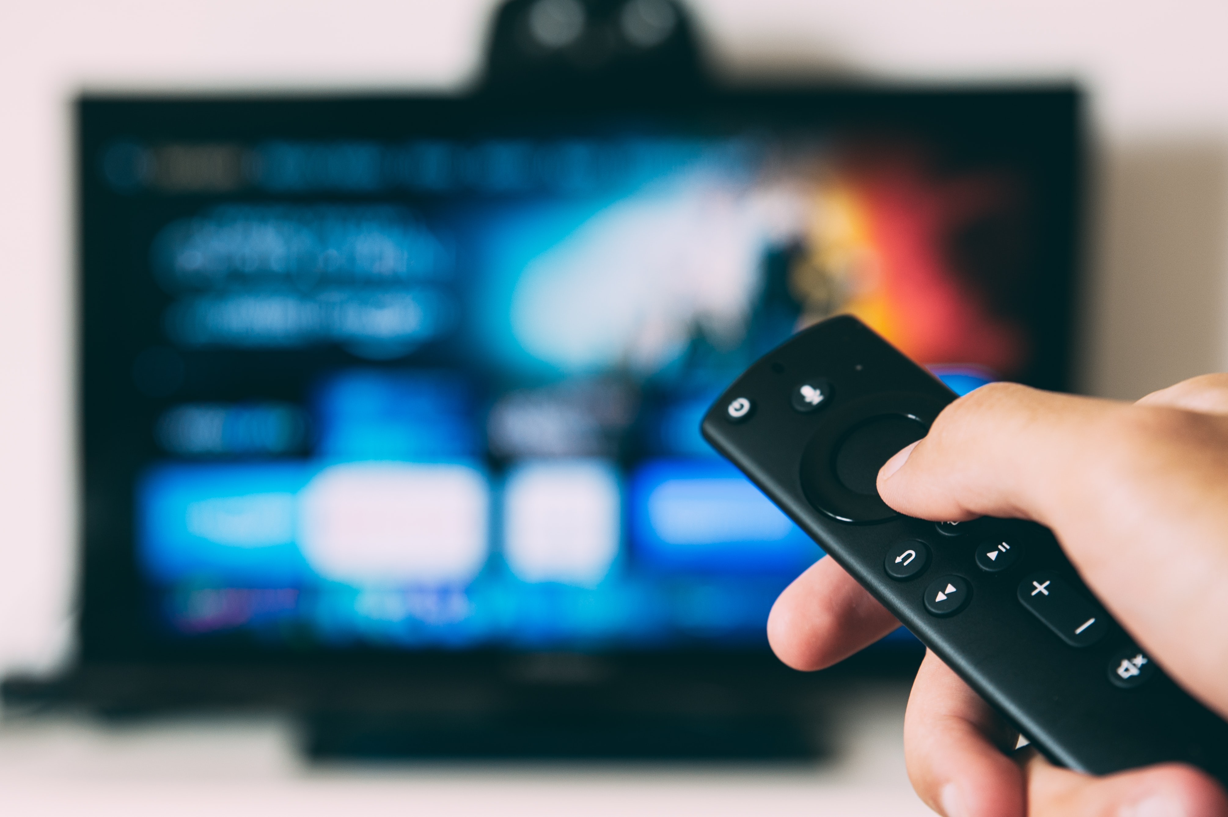 A persons thumb on a TV remote control with a blured out TV in the background.