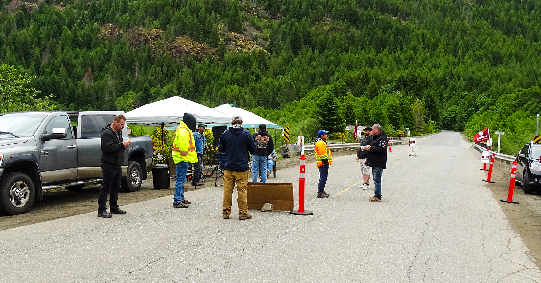A dozen people stand on the road at a remote picket line location with forested mountains in the background